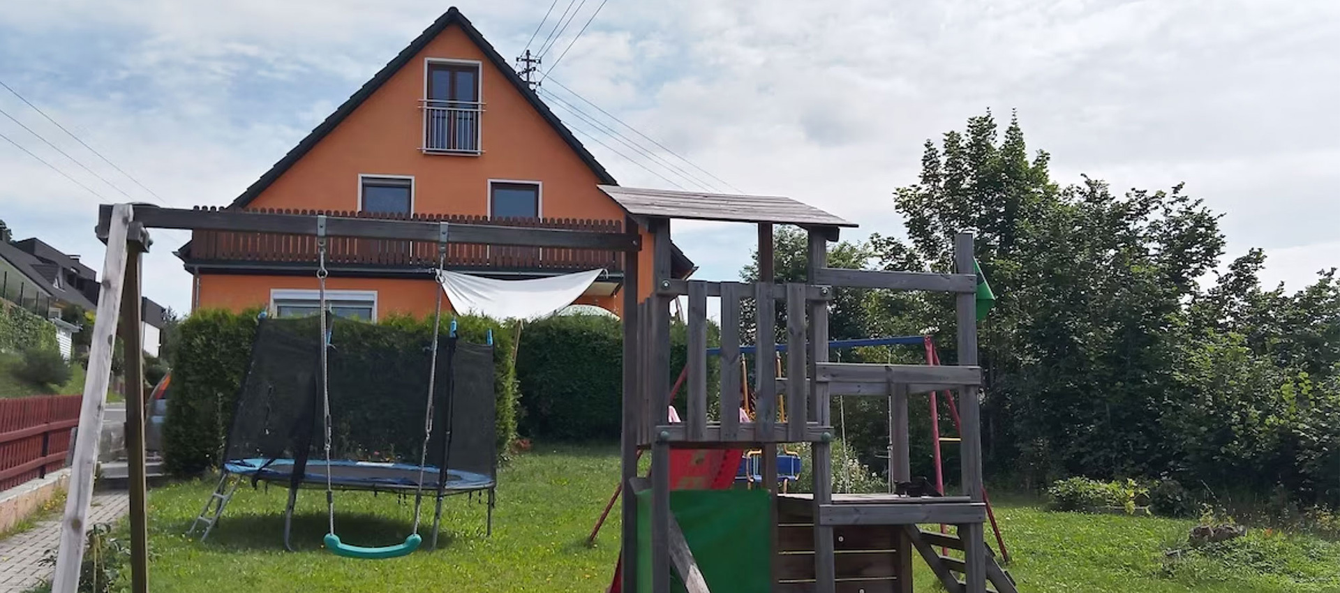 Ein Spielplatz mit Schaukel, Kletterturm und Trampolin. Im Hintergrund ist das Haus zu sehen.
